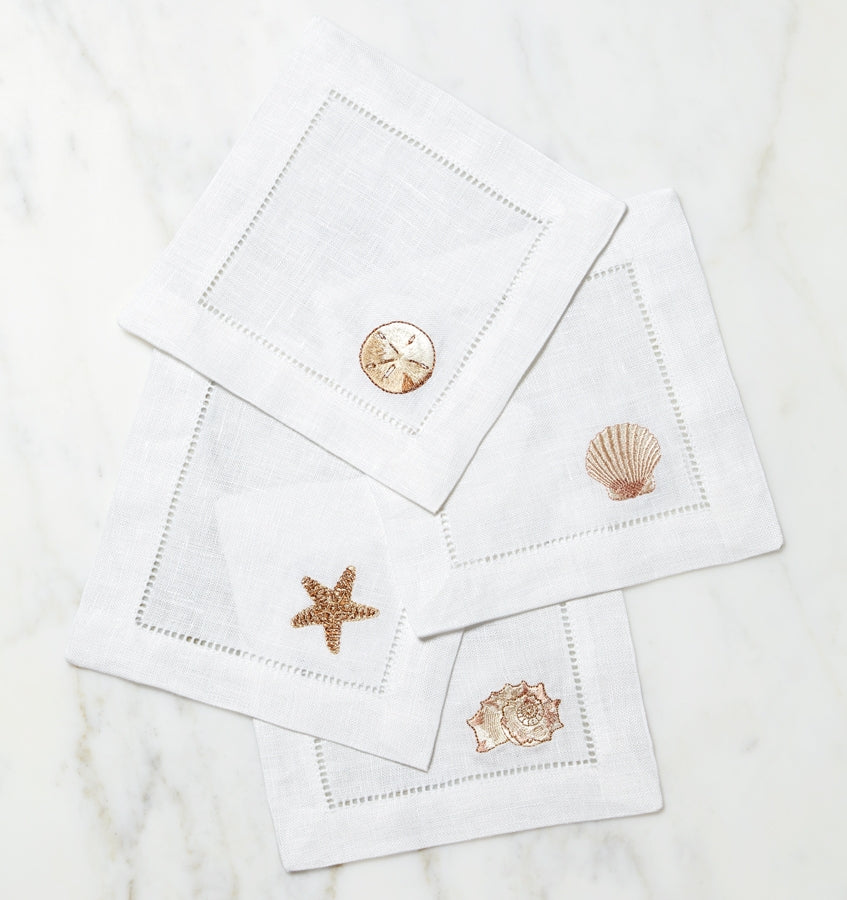 SFERRA Beachcomber cocktail napkins feature four delightful seashell embroideries in golden hues on hemstitched linen napkins.