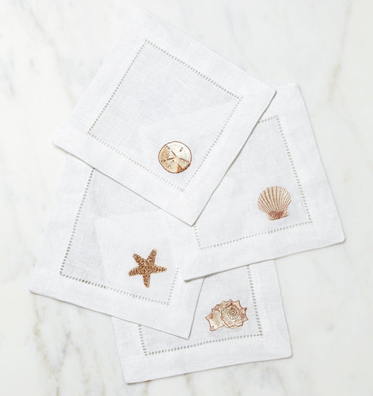 SFERRA Beachcomber cocktail napkins feature four delightful seashell embroideries in golden hues on hemstitched linen napkins.